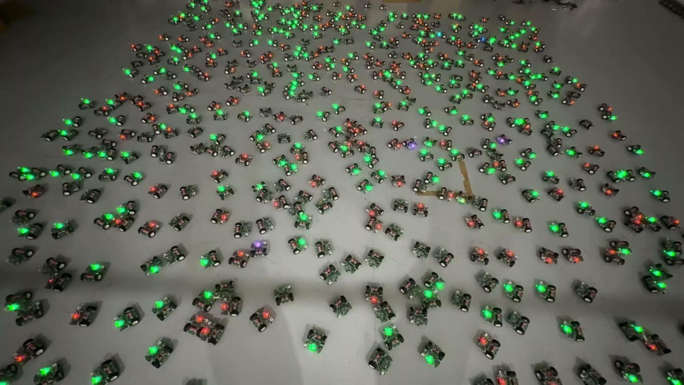 Photo of hundreds of DotBot robots with green LEDs on a grey warehouse floor.